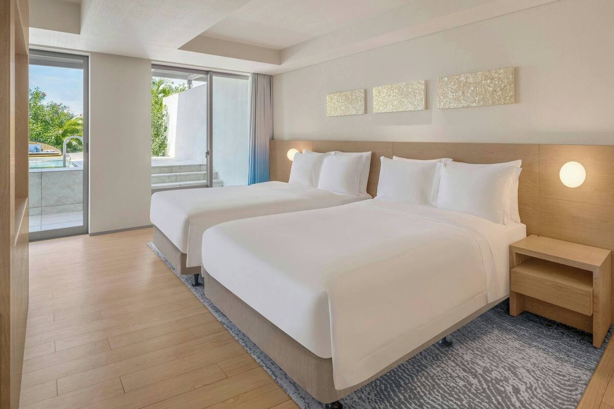 A hotel room with two beds, a wooden headboard, a nightstand, and a view of a swimming pool through a sliding door.