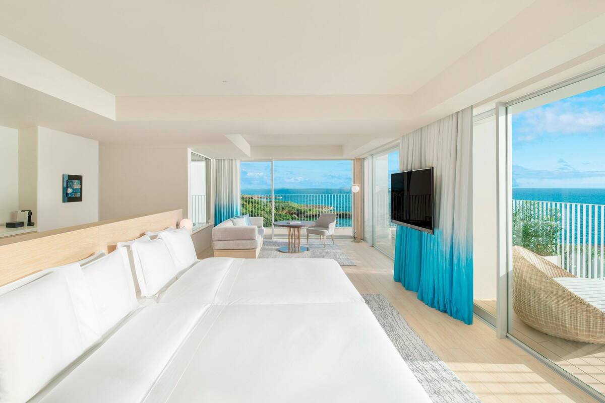 A modern hotel room with a large bed, a TV, a balcony with a view of the sea, and a seating area with a table and chairs.