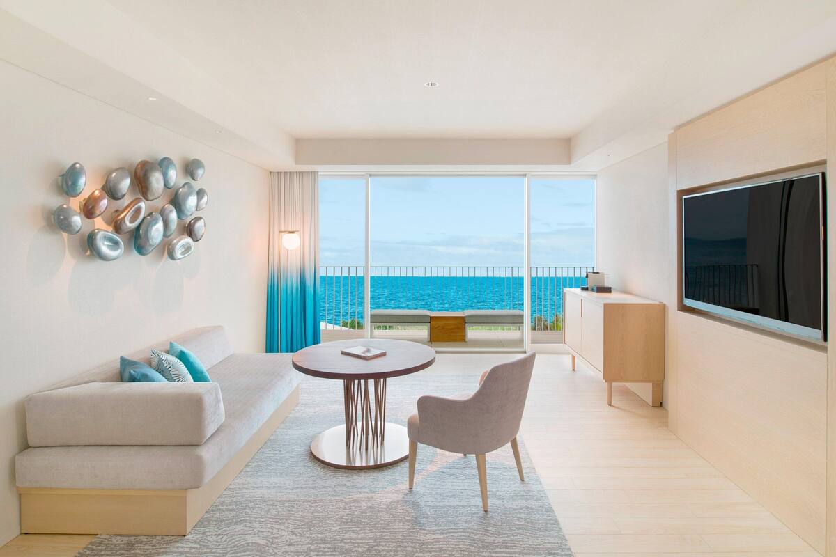 A modern living room with a sofa, a round table, a chair, and a TV. There is a balcony with a view of the sea.