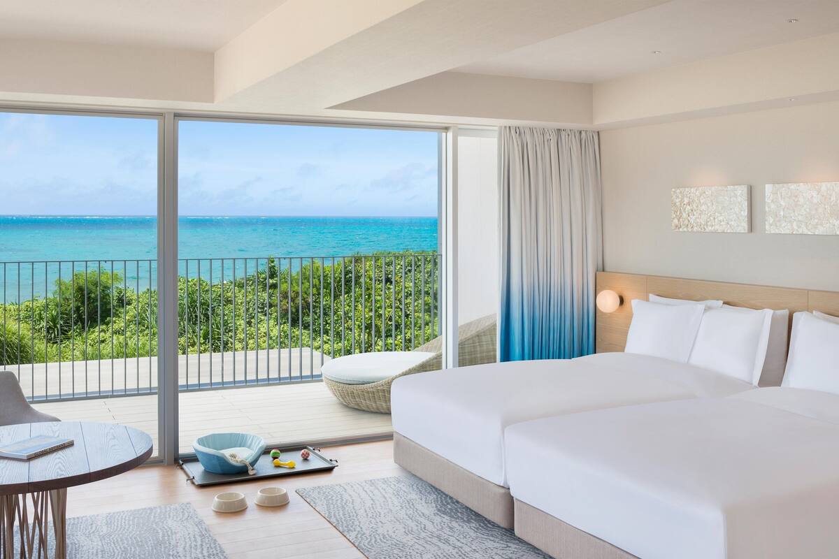 A hotel room with a large balcony, a bed, a seating area, and a view of the ocean.
