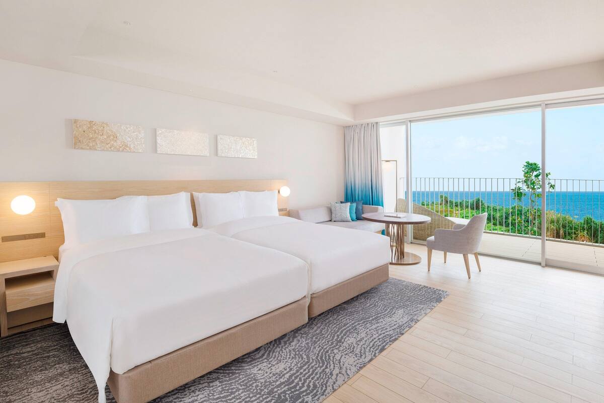 A modern hotel room with a large bed, a small table, and a balcony with a view of the sea.