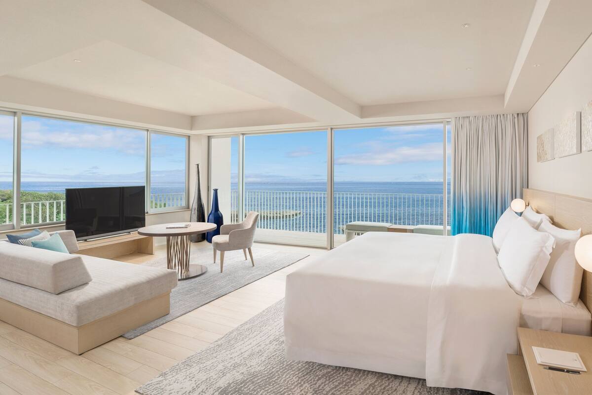 A modern hotel room with a large bed, a sofa, a TV, and a balcony with a view of the sea.