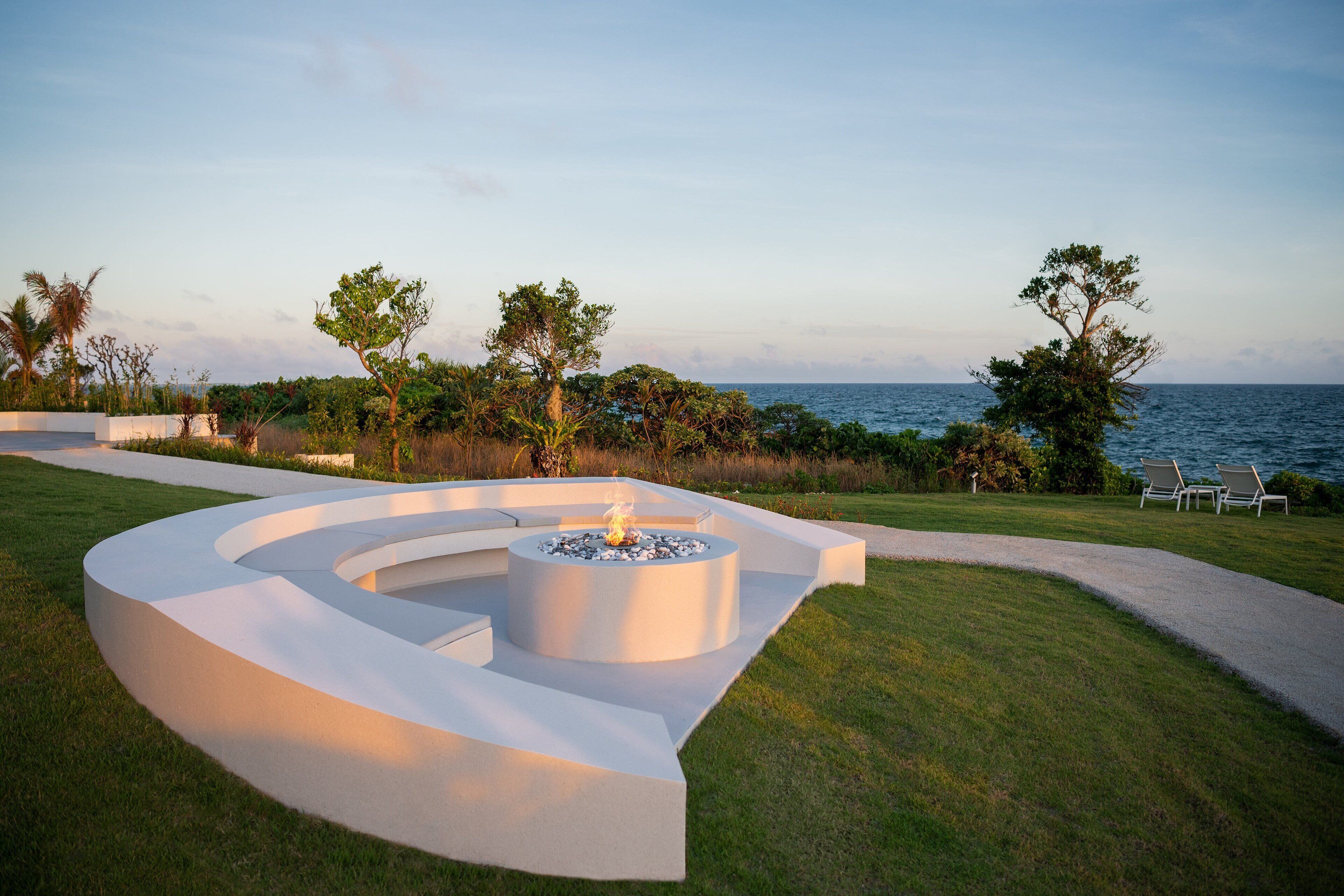 A modern outdoor seating area with a circular fire pit, surrounded by a curved white bench, overlooking a body of water.