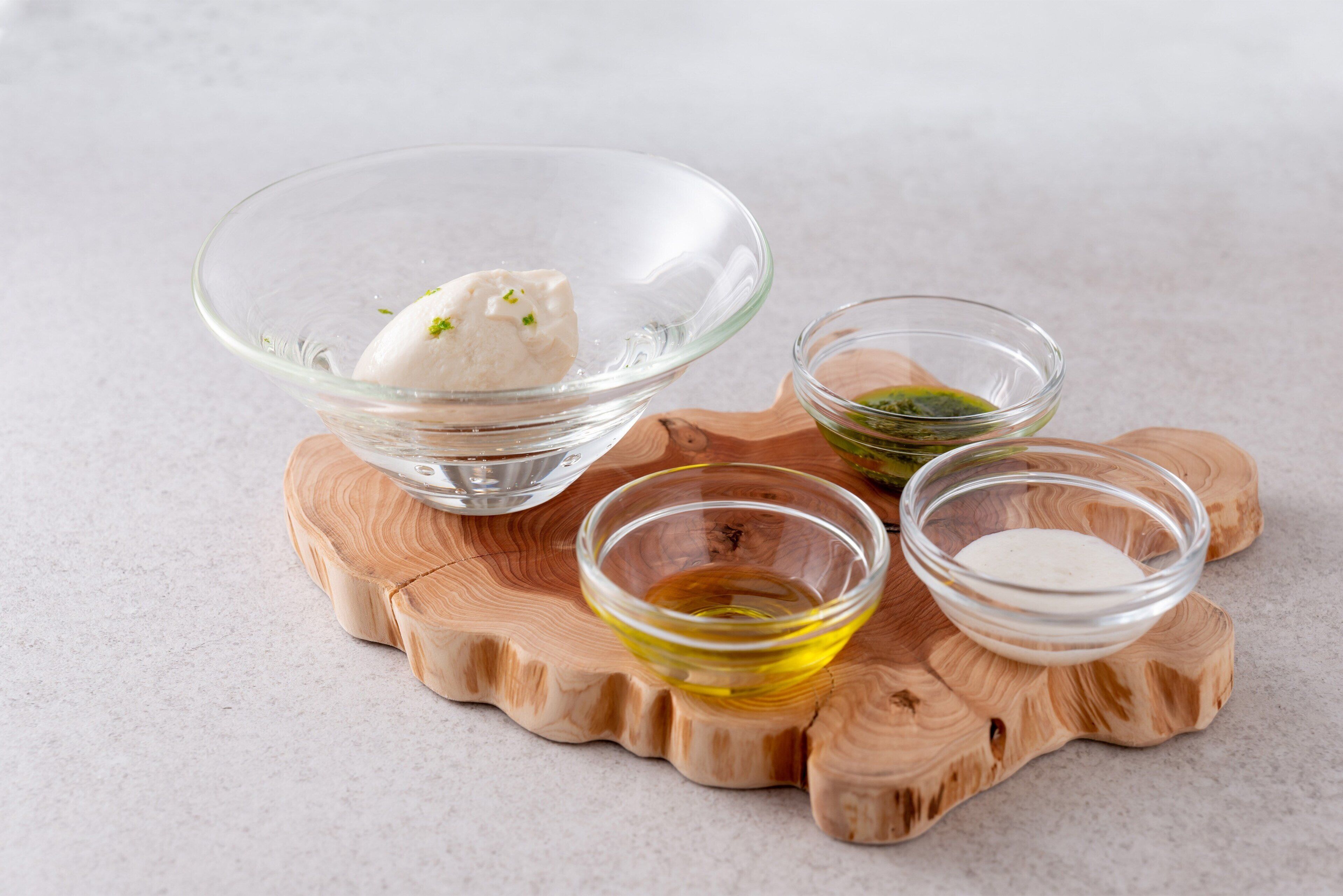 A clear glass bowl with a white substance, two small glass bowls with green and yellow liquids, and a wooden board with a wavy edge.