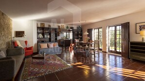 Un salon spacieux comprenant un canapé, un fauteuil et une table basse. On y trouve une bibliothèque remplie de livres et d’objets décoratifs. Un coin repas avec une table et des chaises est visible au fond.