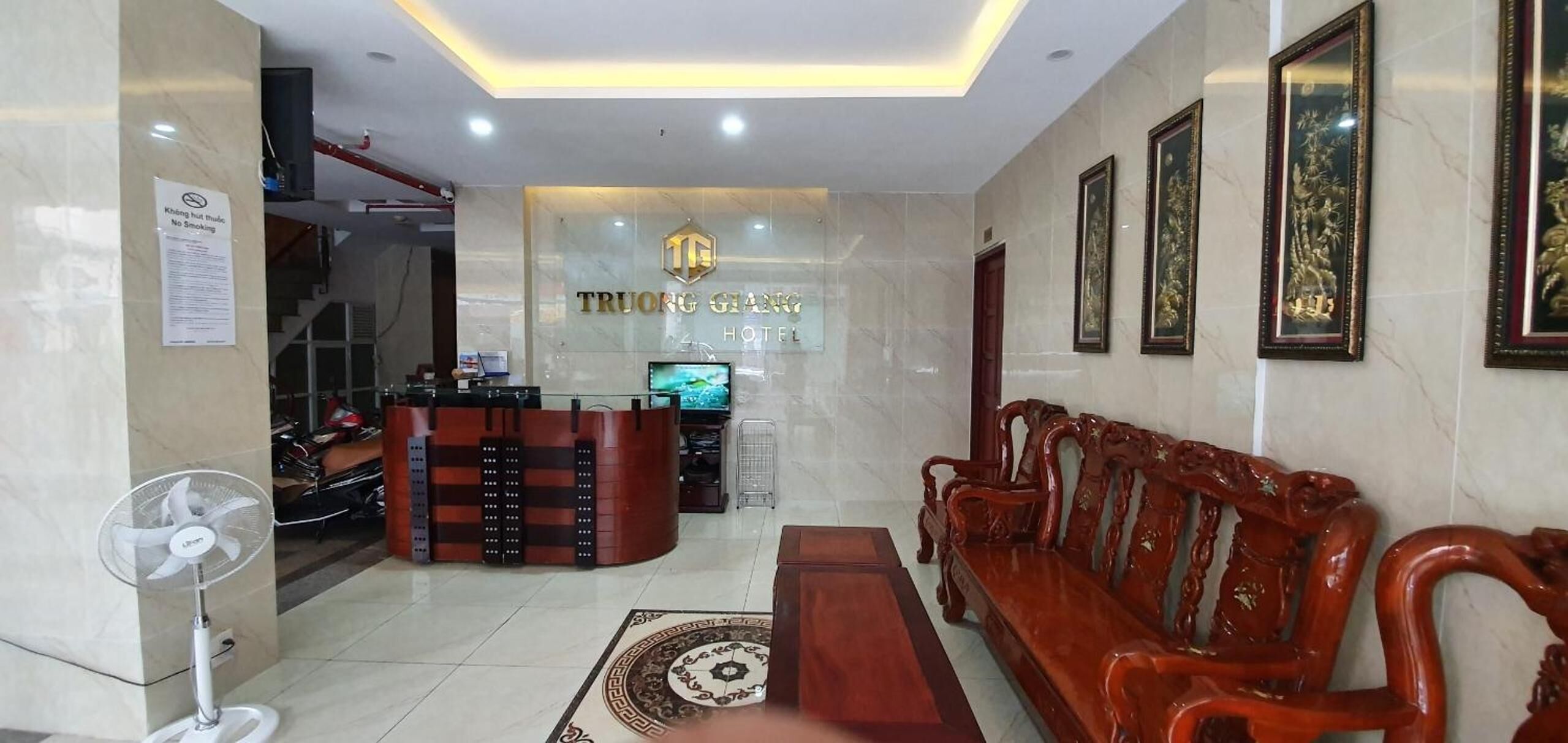 A hotel lobby with a reception desk, a sofa, a fan, and framed artwork on the wall.