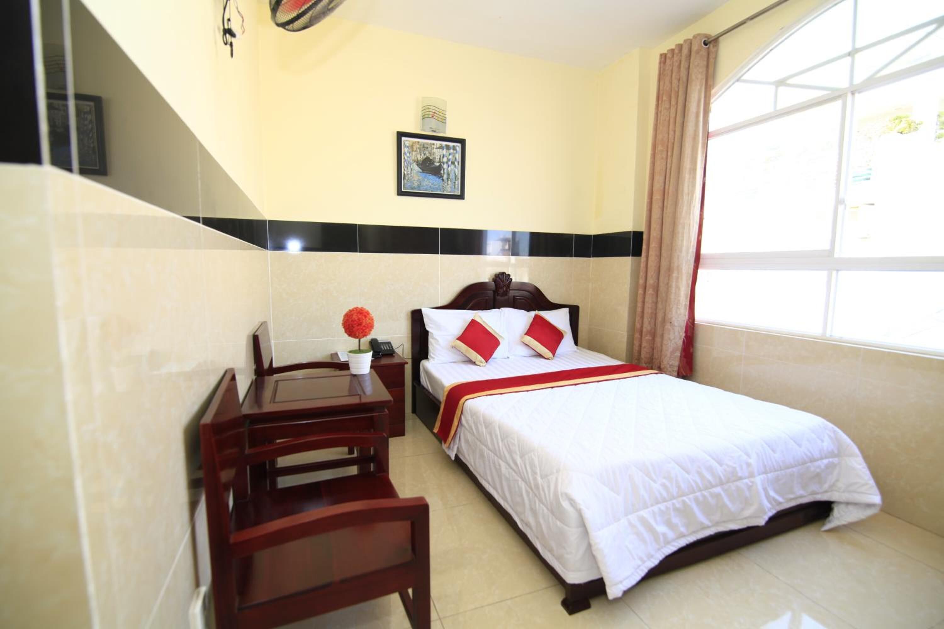Habitación de hotel con cama, escritorio y silla. Hay una ventana con cortinas, un cuadro en la pared y una mesita con un florero.