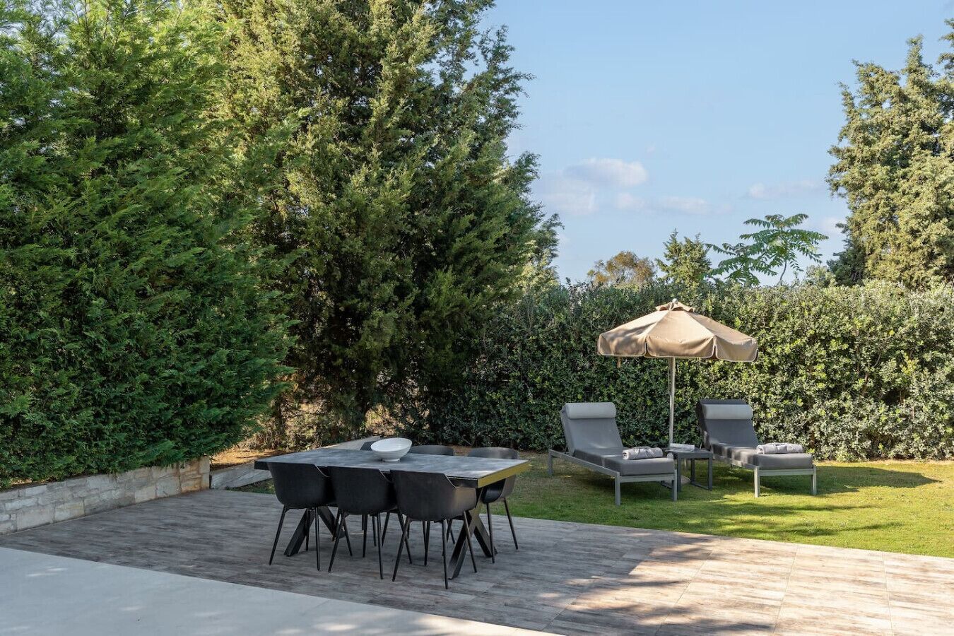 A patio with a table and chairs, an umbrella, and a hedge in the background.