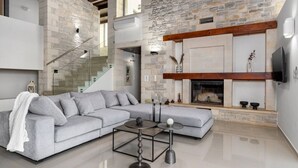 Uma sala de estar moderna com parede de pedra, sofá modular cinza, escada de vidro e lareira.