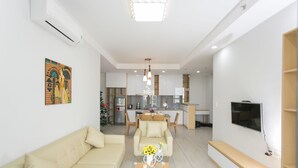 Een moderne woonkamer met een bank, een glazen salontafel met bloemen, een eethoek met stoelen, een keuken op de achtergrond en een tv aan de muur.