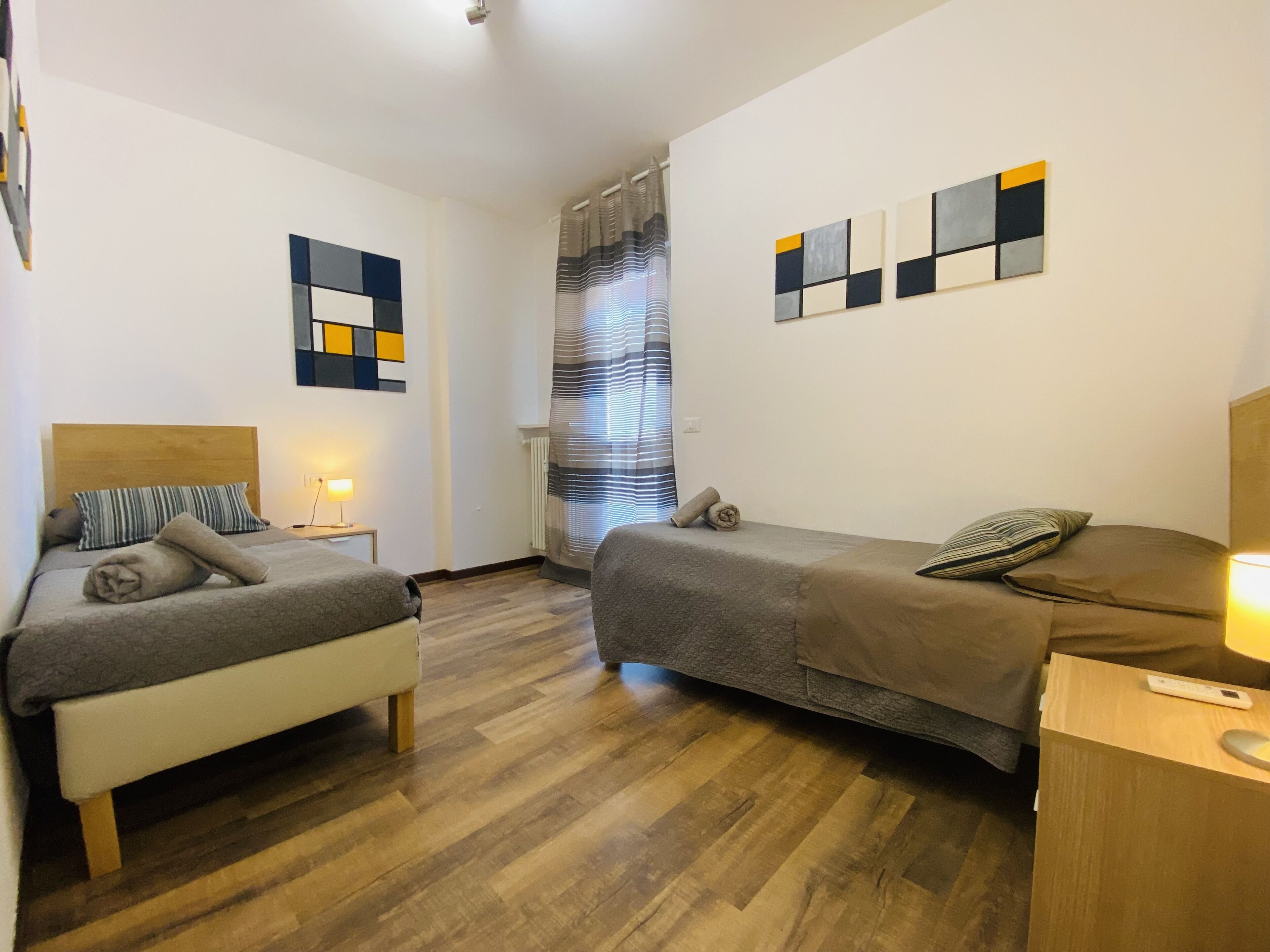 A hotel room with two beds, wooden flooring, a window with striped curtains, and abstract wall art.
