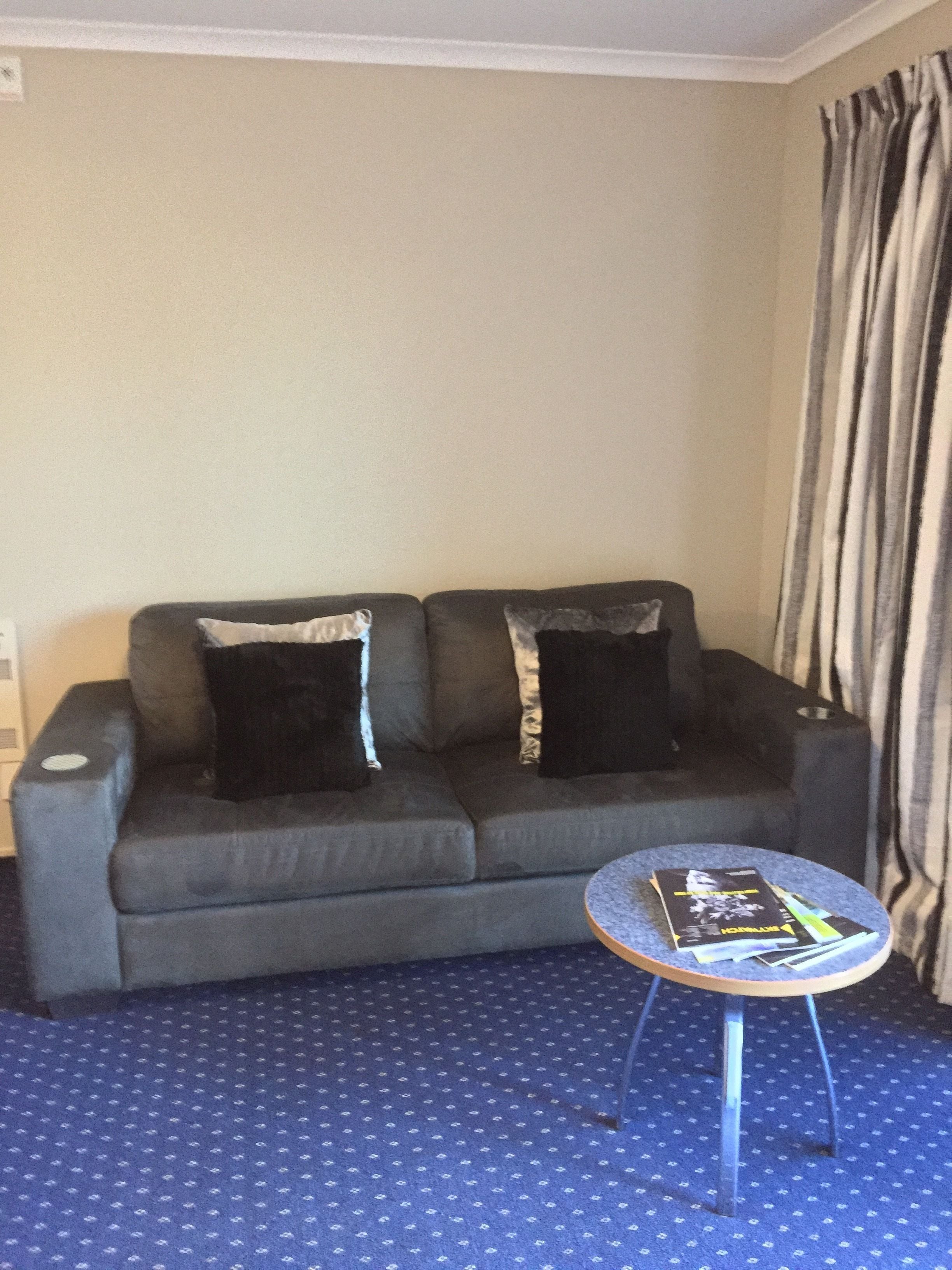 Ein Wohnzimmer mit grauem Sofa, zwei schwarzen Kissen, einem kleinen runden Tisch mit Zeitschriften und einem gemusterten Teppich.