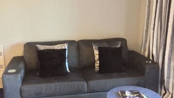 Ein Wohnzimmer mit grauem Sofa, zwei schwarzen Kissen, einem kleinen runden Tisch mit Zeitschriften und einem gemusterten Teppich.
