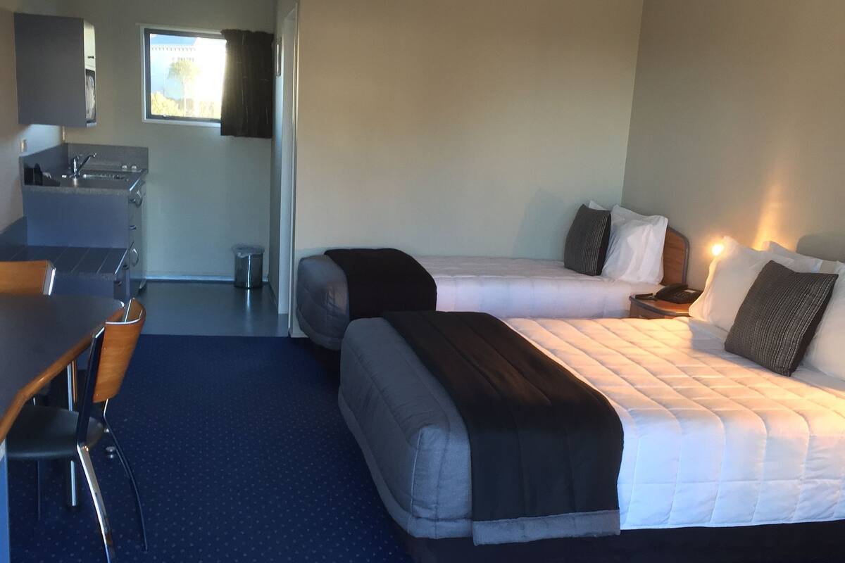 A hotel room with two beds, a desk, a chair, a small kitchenette, and a window.