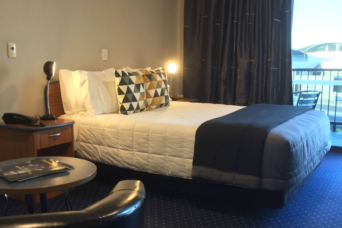 A hotel room with a bed, a desk with a lamp, a chair, and a view of the outside.