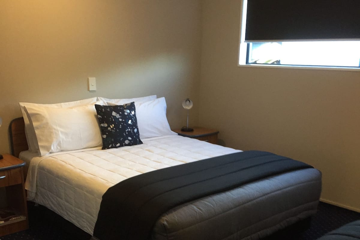 A hotel room with a bed, a nightstand, a lamp, a window, and a chair.
