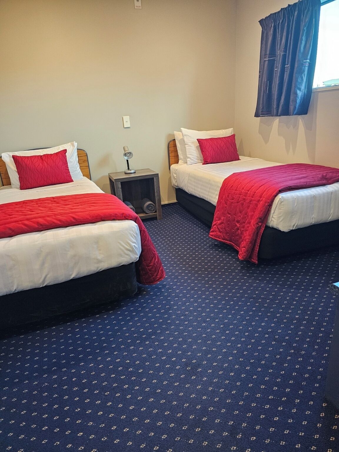 Habitación de hotel con dos camas, cada una con ropa de cama roja y cabeceros de madera. Hay una mesita de noche pequeña con una lámpara, y la habitación tiene una alfombra azul con un diseño discreto.