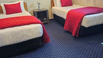 Habitación de hotel con dos camas, cada una con ropa de cama roja y cabeceros de madera. Hay una mesita de noche pequeña con una lámpara, y la habitación tiene una alfombra azul con un diseño discreto.