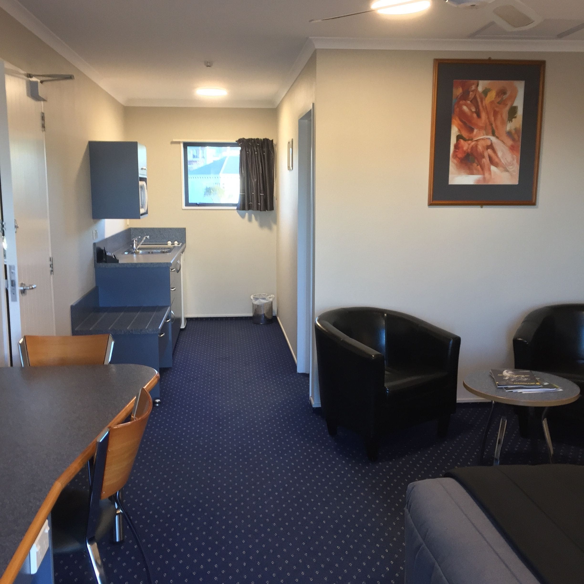 A compact room with a kitchenette, a dining area with a table and chairs, a seating area with a sofa and armchairs, and a framed artwork on the wall.