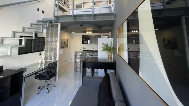 A modern interior with a staircase leading to a loft area, a dining area with a black table and chairs, and a kitchen with a central island.