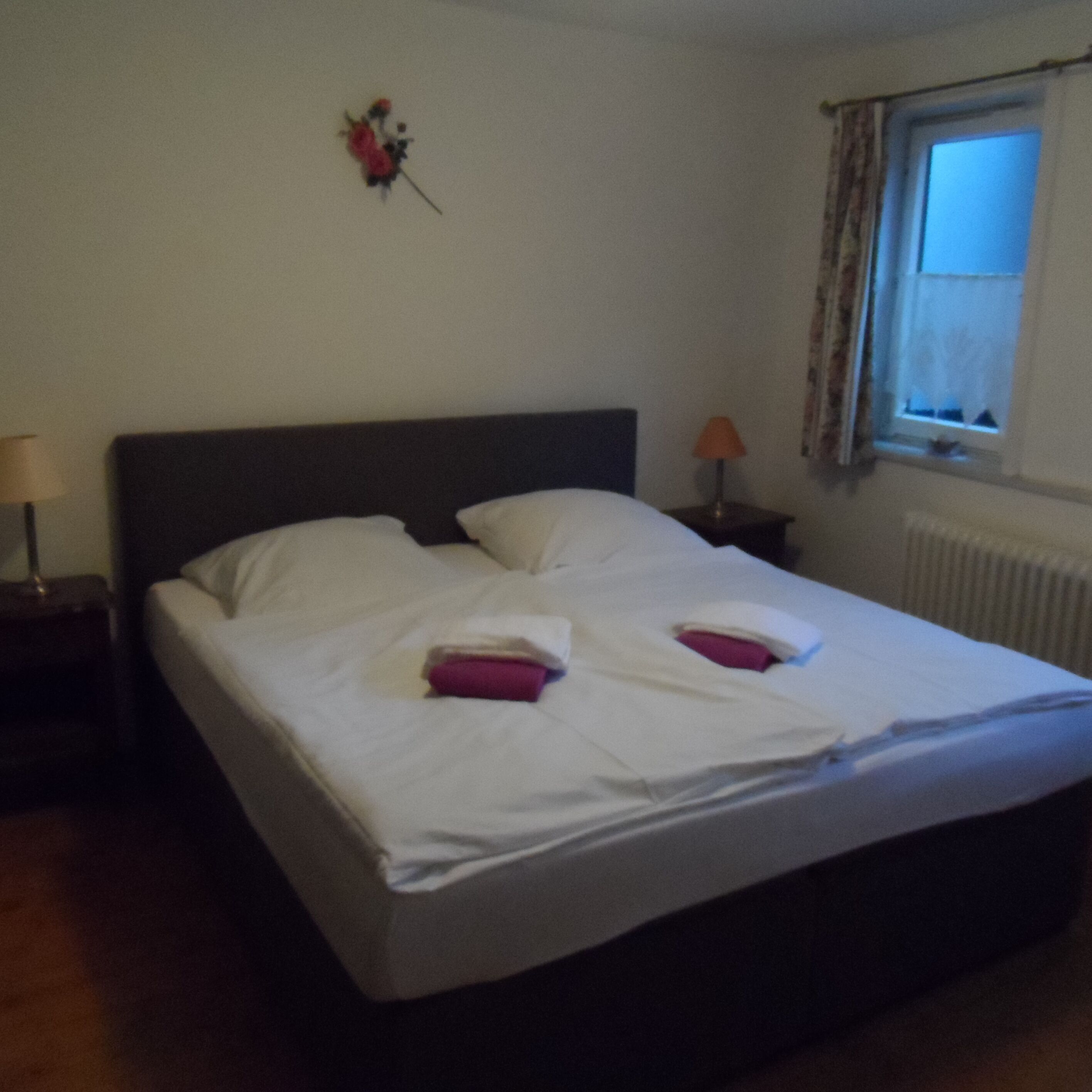A neatly made bed with white linens and two pillows, a bedside lamp, and a window with curtains.