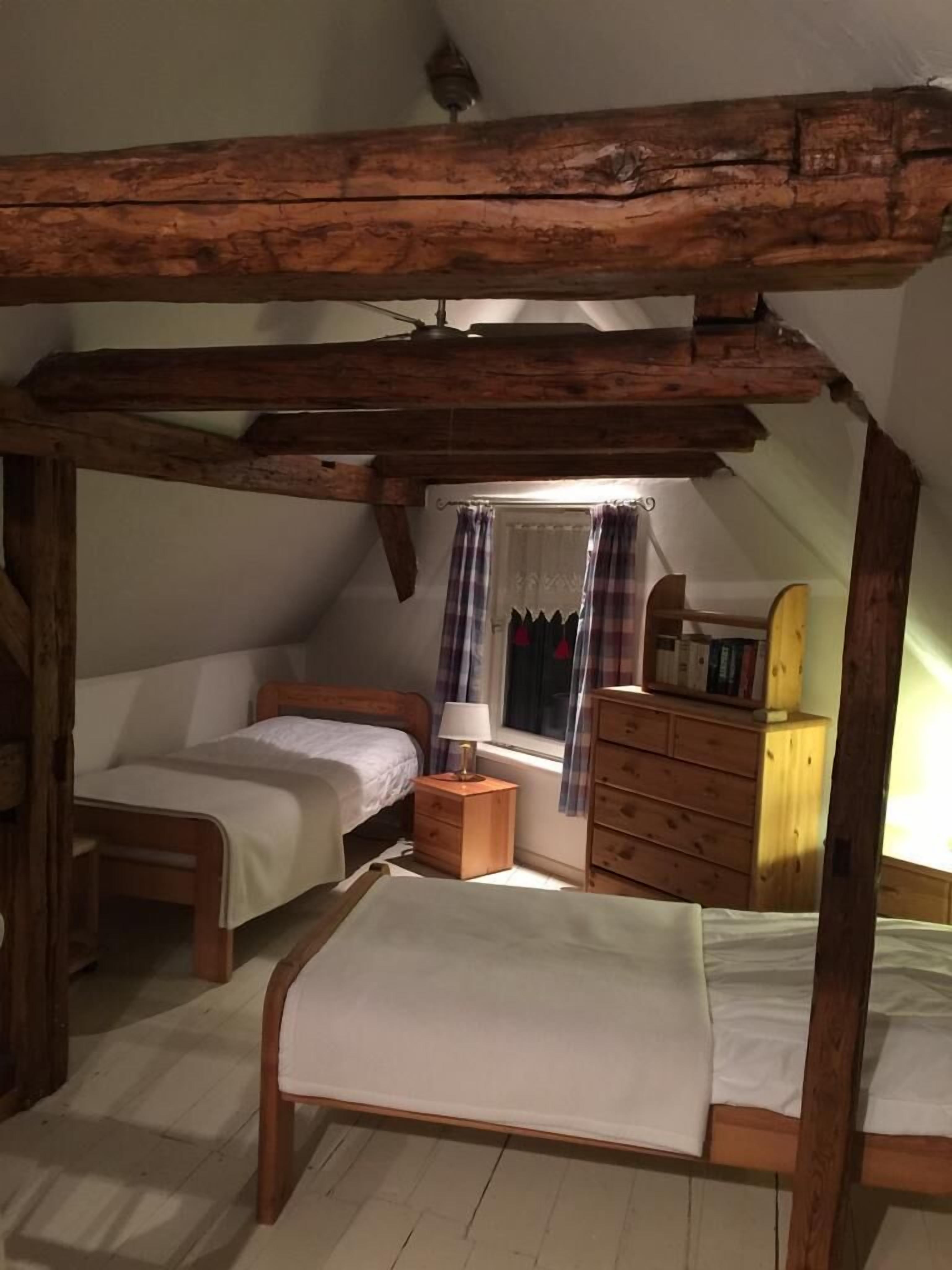 Une chambre confortable située sous les toits, avec des poutres apparentes, deux lits simples, une table de chevet, une commode et une fenêtre avec des rideaux.