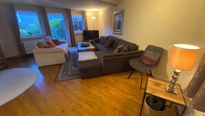 Ein Wohnzimmer mit Sofa, Sessel und einem kleinen Tisch mit Lampe. Ein Fernseher und ein Fenster mit Vorhängen sind ebenfalls vorhanden.