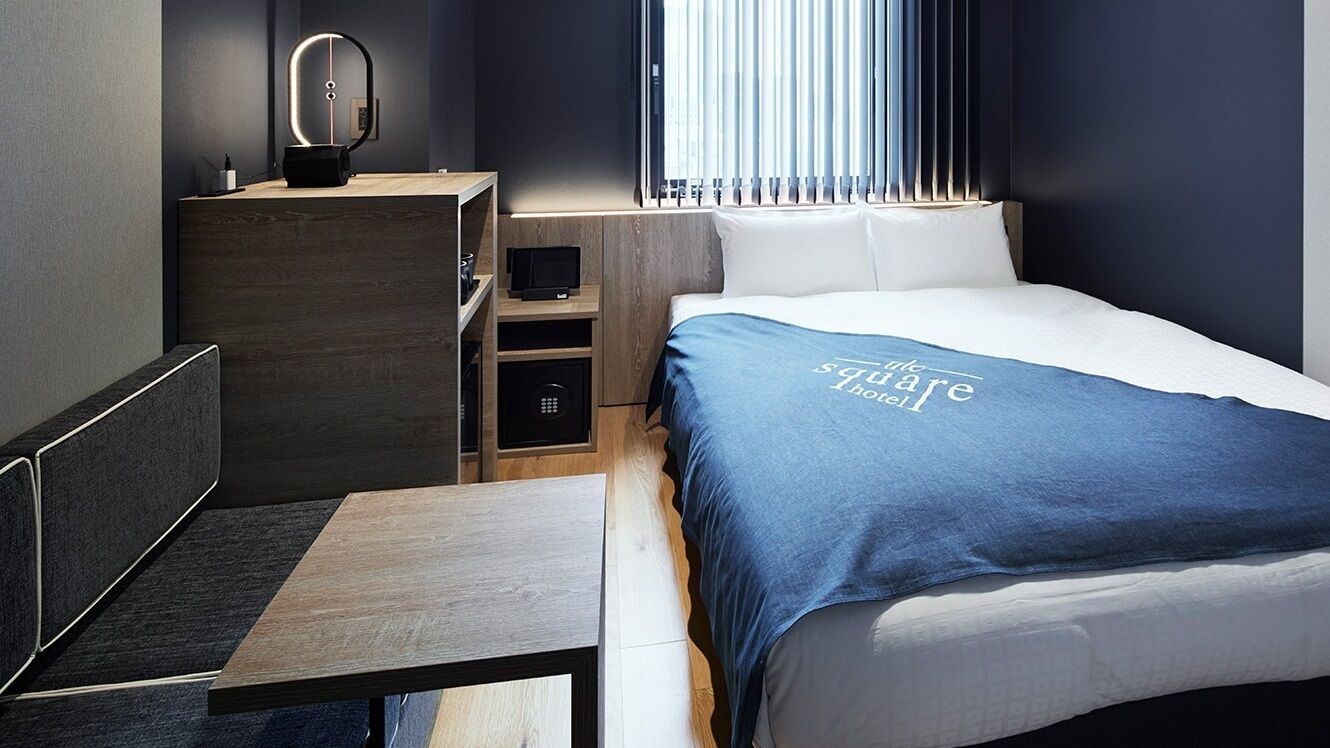 A modern hotel room with a bed, bedside table, and a desk with a chair.