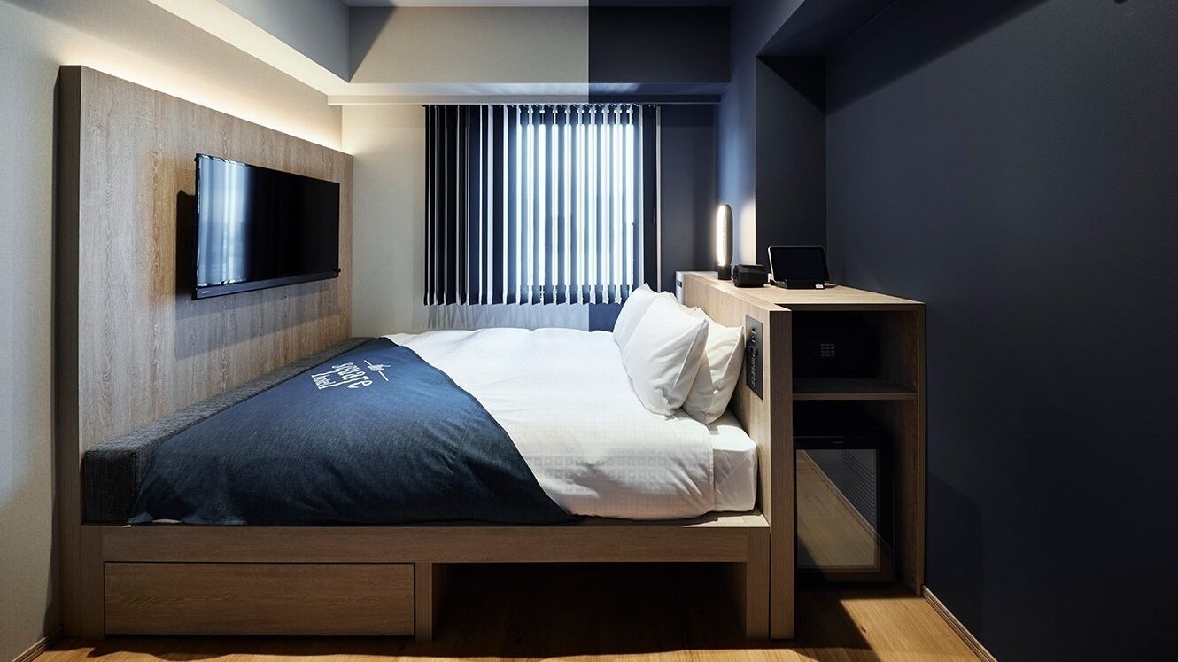 A modern hotel room with a bed, a TV mounted on the wall, a desk with a lamp, and a window with blinds.