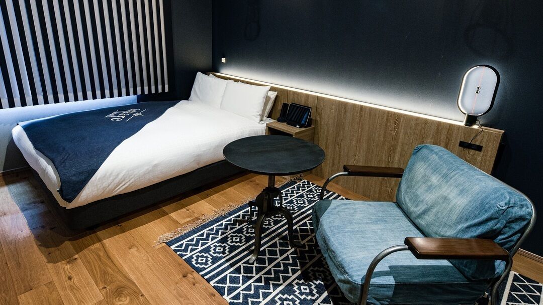 A modern hotel room with a bed, a small round table, a chair, and a bedside table with a lamp.