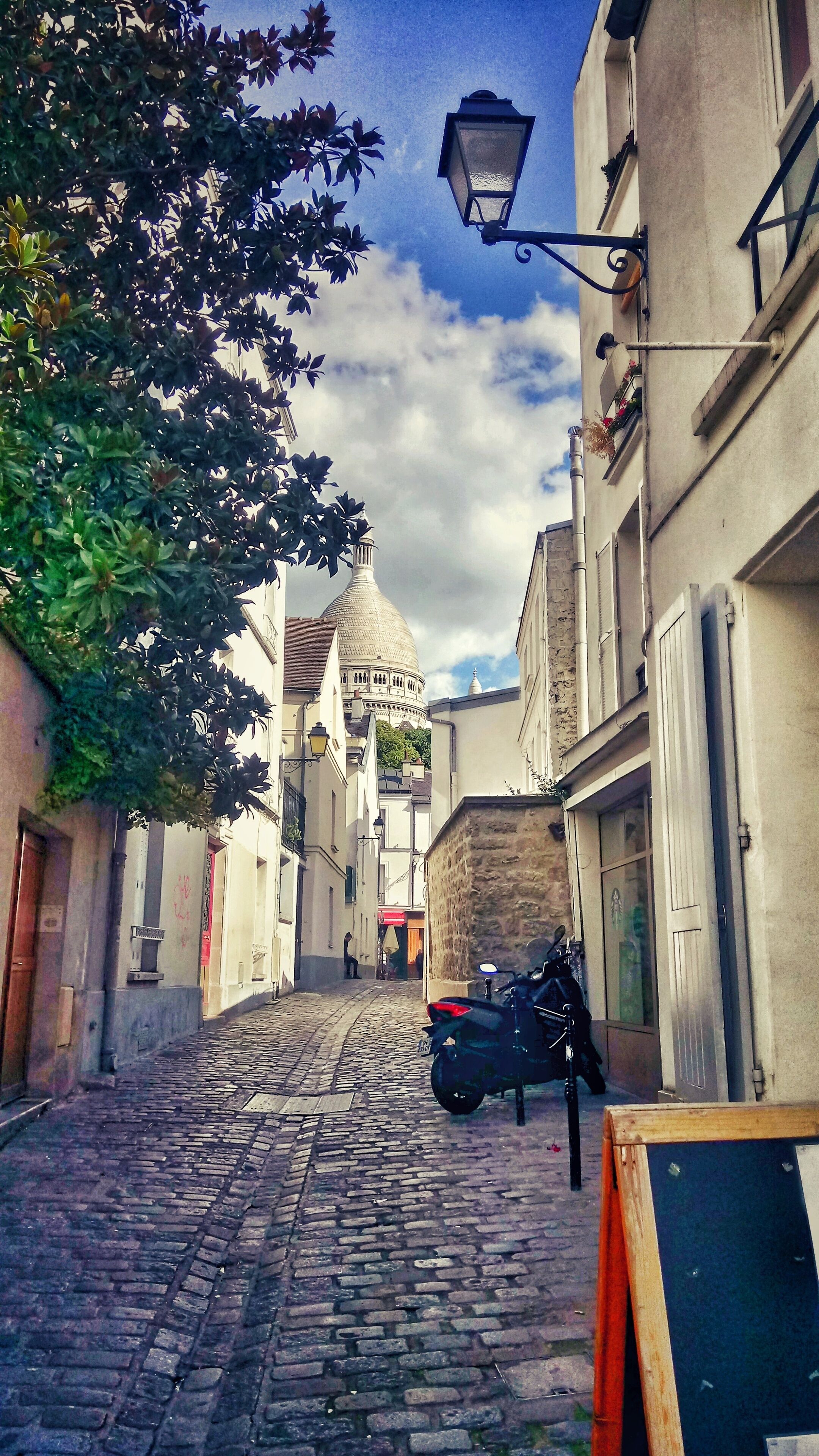 Eine Kopfsteinpflasterstraße mit einem abgestellten Motorrad am Rand, einer Straßenlaterne und einem Gebäude mit Kuppel im Hintergrund.