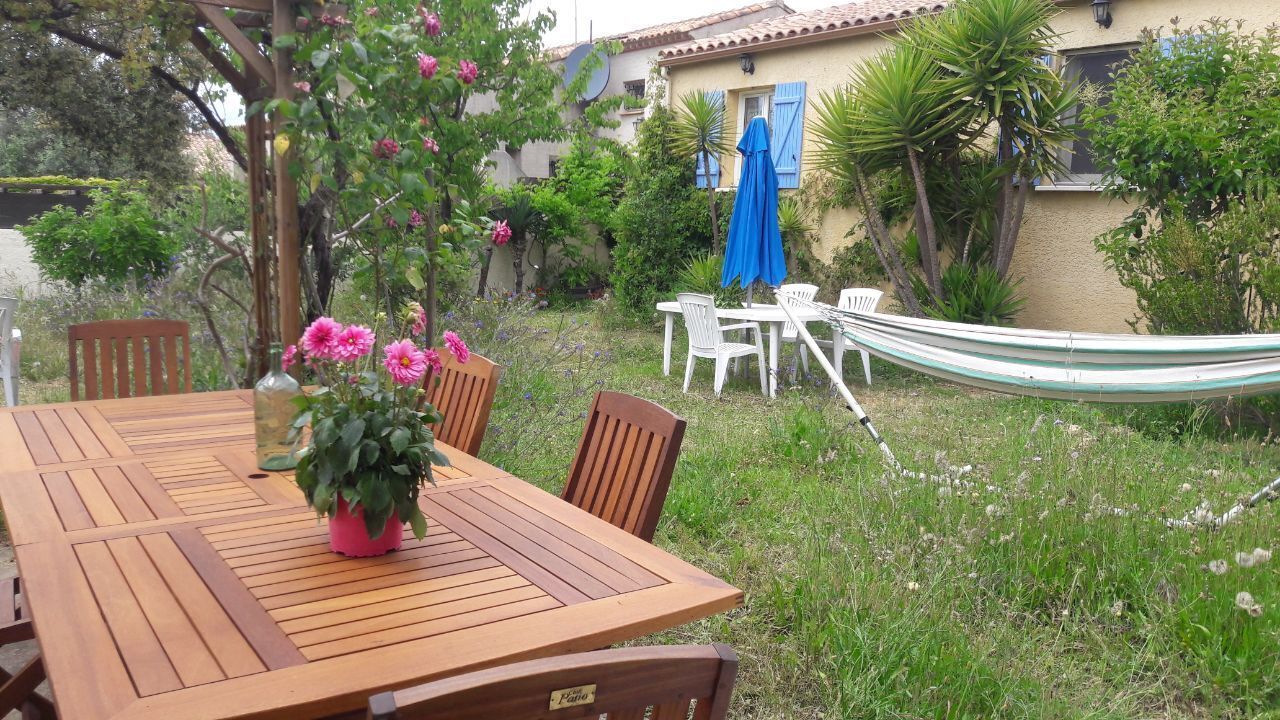 Une table en bois avec des chaises, un hamac et une plante en pot, le tout dans un coin jardin avec une maison en arrière-plan.