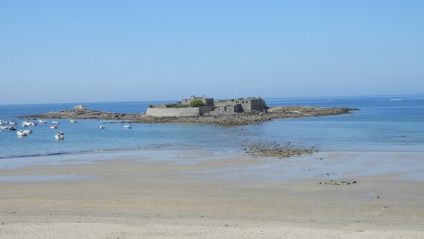 Un paysage côtier avec une petite île rocheuse sur laquelle se trouve un bâtiment, le tout étant entouré d’eaux calmes et de bateaux.