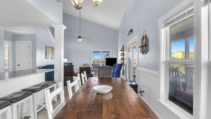 A spacious dining area with a wooden table, white chairs, and a large window with a view of the outdoors. - SeaSide: Oceanview 5 bedroom with easy beach access (Oak Island)