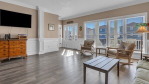 A spacious living room with hardwood flooring, a flat-screen TV, a wooden dresser, and a large window with blinds. - Don't Worry Beach Happy: Ocean View, Swimming Pool (Oak Island)