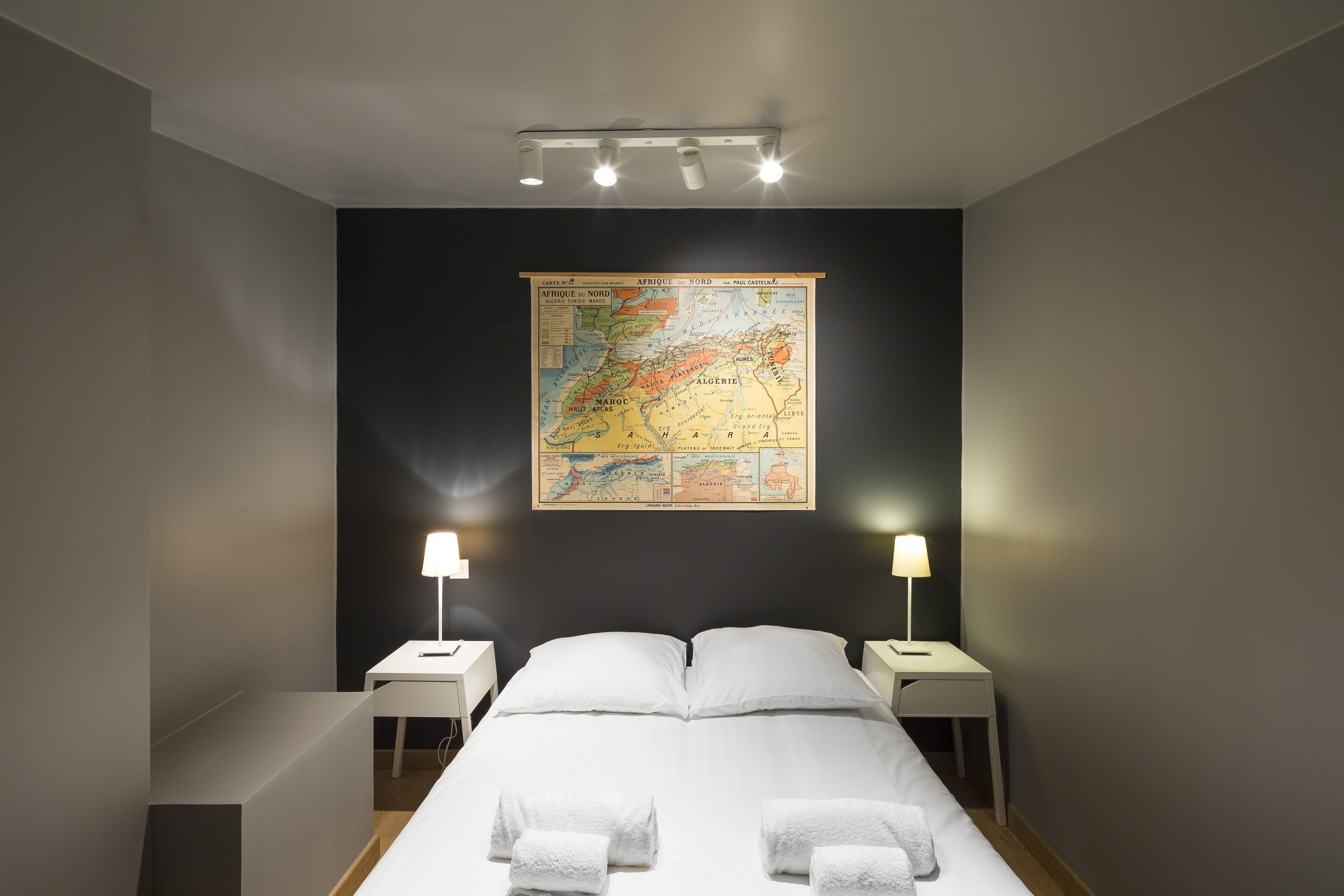 Una camera d'albergo con una grande mappa appesa al muro, un letto con lenzuola bianche, due comodini con lampade e un moderno lampadario.
