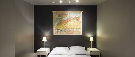 Una camera d'albergo con una grande mappa appesa al muro, un letto con lenzuola bianche, due comodini con lampade e un moderno lampadario.