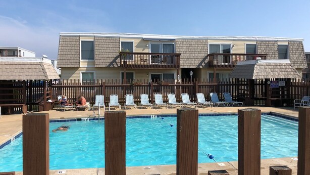 A pool area with a wooden fence, lounge chairs, and a building with balconies. - Beautiful 2-BR Bayside Condo W/Pool, Gull Way Villas, Short Walk To Beach (Ocean City)