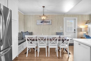 A modern kitchen with white cabinetry, a stainless steel refrigerator, a dining table with white chairs, and a wooden wall with a fish painting. - The Beachcomber: Spacious, Family-friendly, Dog-friendly, Oceanfront Home with G (Oak Island)