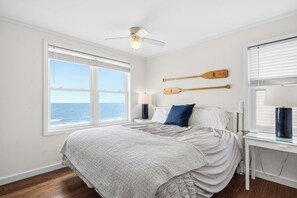 A bedroom with a large window, a bed, a ceiling fan, and two wooden paddles on the wall. - The Beachcomber: Spacious, Family-friendly, Dog-friendly, Oceanfront Home with G (Oak Island)