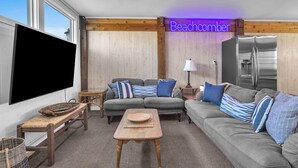 Uma sala de estar com um sofá cinzento, uma mesa de centro de madeira, uma televisão de tela plana e um letreiro neon com a inscrição "Beachcomber".