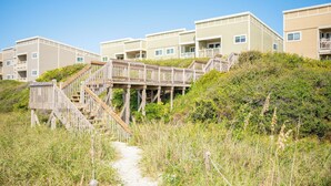 Een strandhotel met meerlaags gebouwen, houten trappen die naar balkons leiden, en een zandpad.