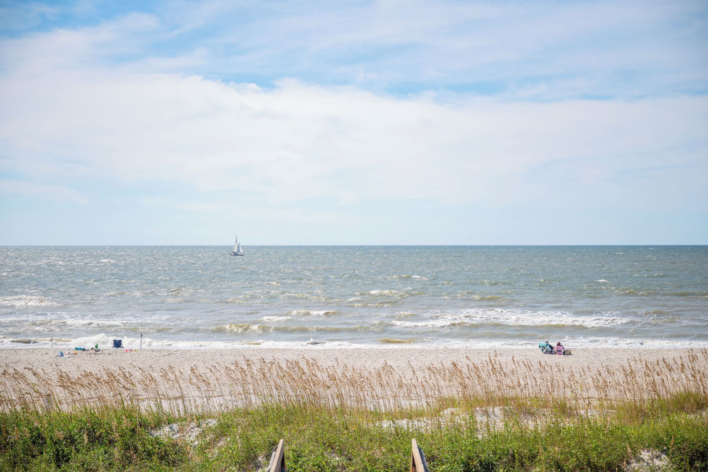 Een strand met een zeilboot in de verte, een zandstrand en wat begroeiing op de voorgrond.