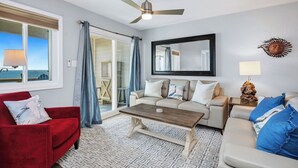 Een woonkamer met een rode fauteuil, een beige bank en een salontafel. Er is een plafondventilator, een grote spiegel en een uitzicht door een glazen deur.