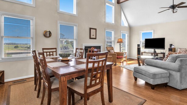 A spacious living room with a dining area, large windows, and a ceiling fan. - Heron Pointe: Oceanwalk home with waterway views and just a short stroll to the (Oak Island)