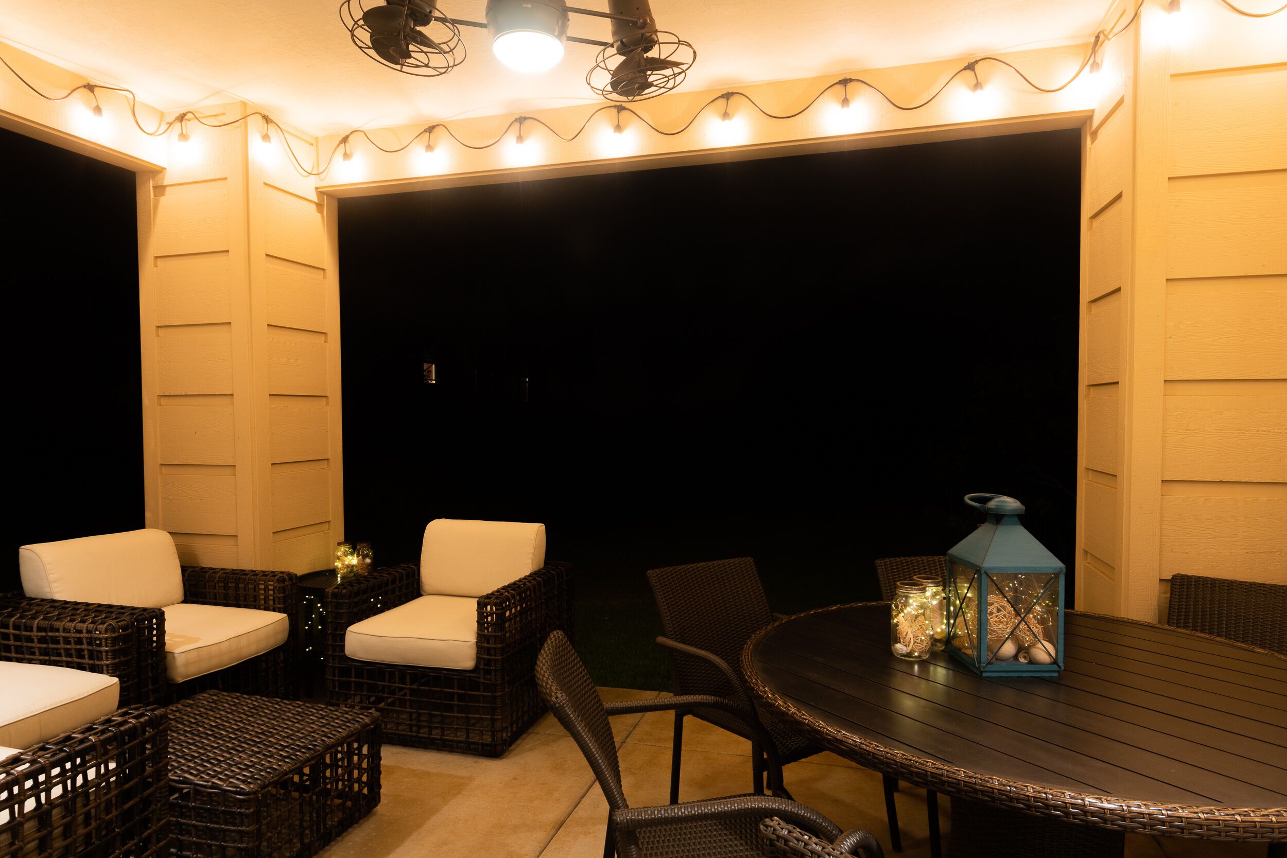 Un espace extérieur aménagé en terrasse, avec des meubles en osier et une table, agrémenté de guirlandes lumineuses et d’une lanterne.