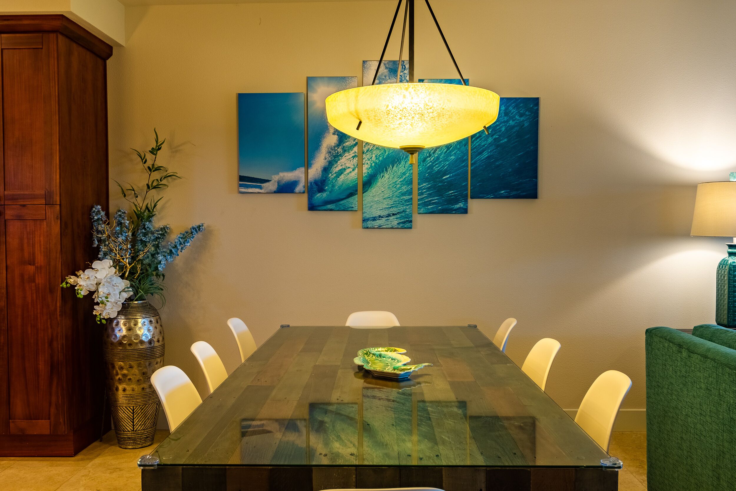 A dining area with a glass table, white chairs, a green sofa, and a decorative vase with flowers.