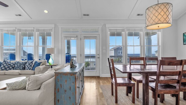 A spacious living room with a large window, a dining area with wooden chairs, and a rustic blue cabinet. - Coast is Clear: Expansive Ocean and Waterway Views w/Private Backyard Pool (Oak Island)