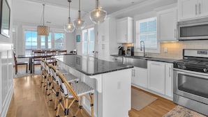 A modern kitchen with white cabinetry, a central island with bar stools, stainless steel appliances, and pendant lighting. - Coast is Clear: Expansive Ocean and Waterway Views w/Private Backyard Pool (Oak Island)