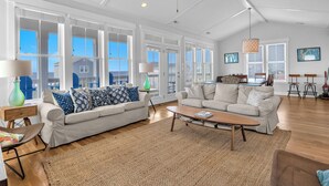 A spacious living room with two sofas, a coffee table, and a ceiling fan. Large windows provide ample natural light and views of the outside. - Coast is Clear: Expansive Ocean and Waterway Views w/Private Backyard Pool (Oak Island)